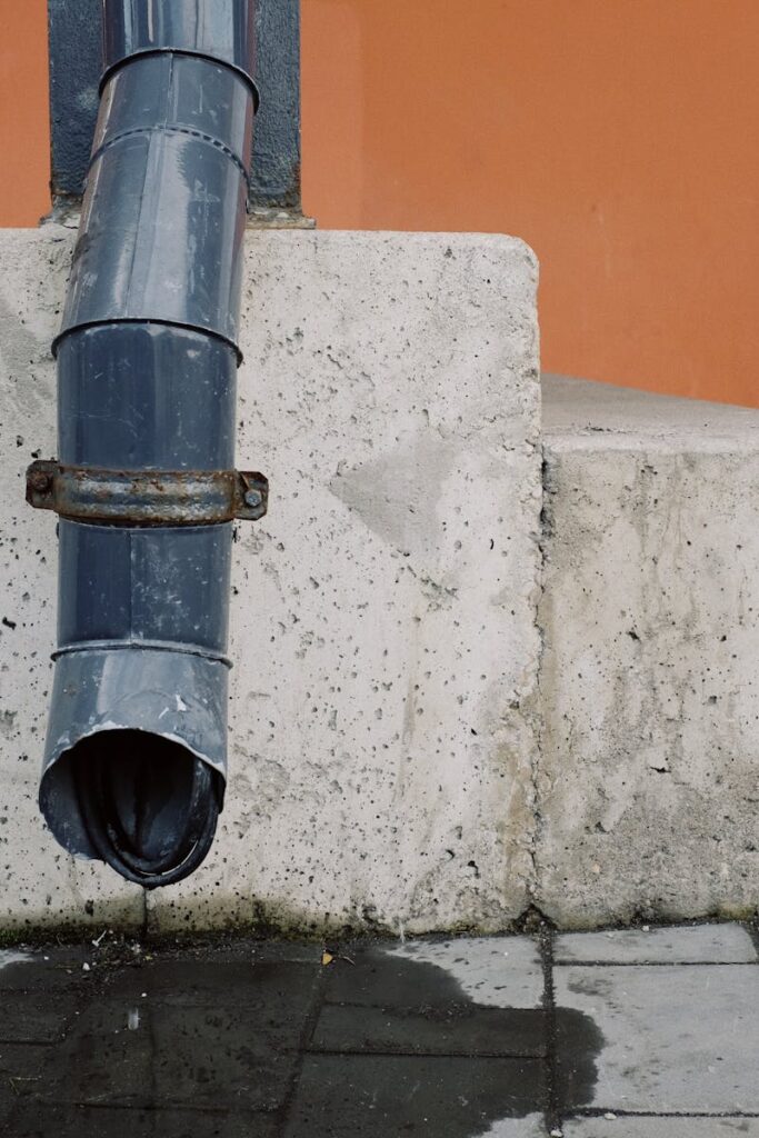 Metal drain pipe with water drops located on red wall near concrete stairs of modern building on street in city